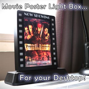 May include: A black movie poster light box with a film strip border. The box is displaying a poster for the movie Pirates of the Caribbean: The Curse of the Black Pearl. The box is on a black base and says "Now Showing" and "For your Desktop!"