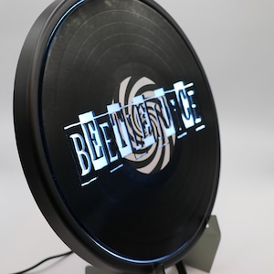 May include: A black vinyl record with a white and black spiral design. The words "BEETLEJUICE" are printed in white on the record.