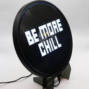 May include: A black circular sign with a white border and a blue light behind it. The sign says "BE MORE CHILL" in white block letters. The sign is on a black stand.