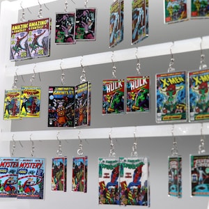 May include: Comic book-themed earrings displayed on a white rack. The earrings feature various comic book covers, including "Amazing Fantasy," "Hulk," and "X-Men." The earrings are rectangular and hang from silver hooks, showcasing vibrant colors and detailed artwork.