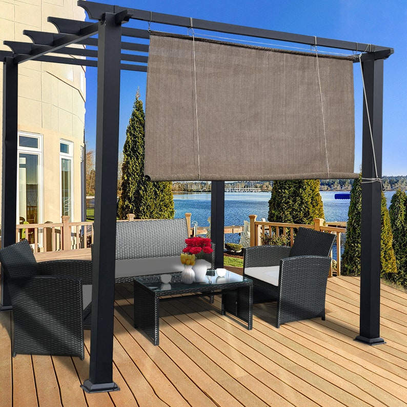 May include: A black pergola with a brown shade, a glass-top coffee table, and black wicker furniture. The outdoor setting includes a wooden deck, a lake, and a house in the background. The scene is set on a sunny day.