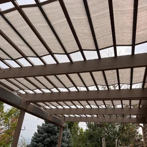 May include: A brown wooden pergola structure featuring a series of horizontal beams and vertical posts. A light beige, semi-transparent fabric shade is draped across the top of the wooden frame, providing partial coverage and filtering sunlight.