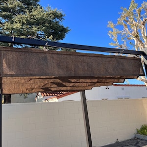 May include: A black metal pergola with a brown fabric shade. The pergola is attached to a white brick wall.