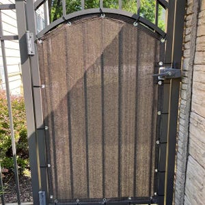 May include: A brown and black privacy screen mesh attached to a dark metal arched gate. The rectangular fabric features vertical stripes, reinforced metal grommets along the edges, and is secured to the gate frame with black ties.