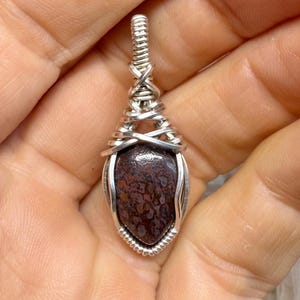 May include: A handcrafted pendant featuring a teardrop-shaped stone with a mottled pattern of brown and red hues. The stone is encased in a silver wire wrap with intricate woven detailing, creating a unique and elegant piece of jewelry.