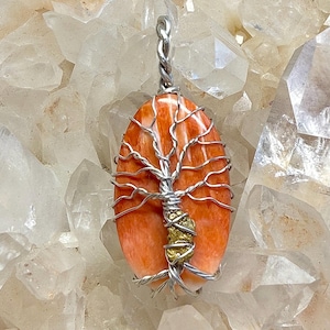 Può includere: Ciondolo ovale in pietra preziosa arancione con un design ad albero in filo d'argento. Il ciondolo presenta un'ansa argentata attorcigliata e un piccolo accento color oro. La pietra preziosa è lunga circa 5 cm.