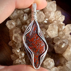 May include: A handcrafted pendant featuring a red and black patterned stone encased in a silver wire wrap. The pendant has a diamond shape and is suspended from a silver bail. The background is a cluster of clear crystals.