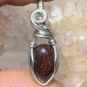 May include: A handcrafted pendant featuring a polished, oval-shaped gemstone with a dark brown and orange pattern. The stone is encased in a silver wire wrap with a decorative swirl at the top. The pendant is suspended from a silver bail.