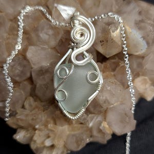 Op de afbeelding: Een zilveren draadgewikkelde hanger met een lichtblauwe zeeglassteen. De hanger hangt aan een zilveren ketting.