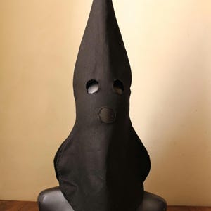 May include: A black pointed hood with eye and mouth holes. The hood is made of fabric and is worn on a black mannequin.