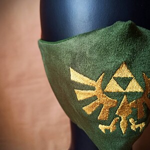 May include: A green face mask with a gold embroidered Triforce symbol from the Legend of Zelda video game series.
