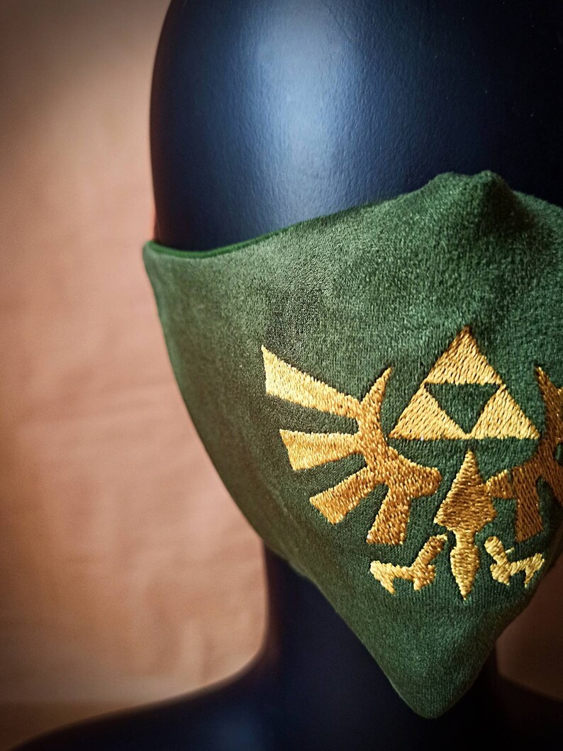 May include: A green face mask with a gold embroidered Triforce symbol from the Legend of Zelda video game series.