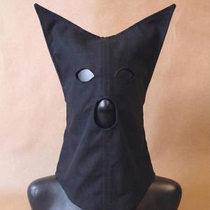 May include: A black fabric hood with eye and mouth openings, displayed on a mannequin head. The hood has pointed ears and a neck covering. The background is a solid tan color.