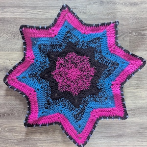 May include: A handmade, star-shaped crochet item in shades of pink, blue, and black. The design features concentric layers of color, with a beaded edge. The item is likely a decorative piece or a small mat.