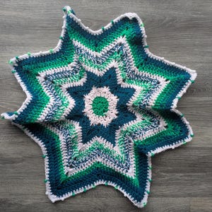 May include: A star-shaped crochet blanket in teal, green, and white. The blanket has a detailed, layered design with a white border. The soft, textured yarn creates a cosy and decorative item. The blanket is approximately 70 cm in diameter.
