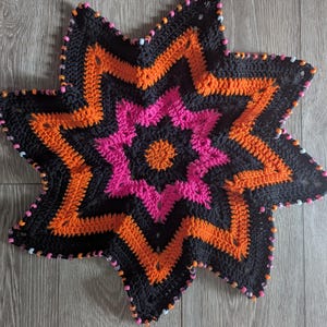 May include: A handmade, star-shaped crochet item in black, orange, and pink. The star has a central pink flower design, surrounded by orange and black zig-zag patterns. The edges are adorned with small, colorful beads.