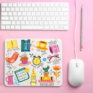 May include: A white computer keyboard, mouse, and a pencil with silver glitter are on a pink surface. A white mousepad features colorful illustrations of books, a teacup, glasses, and the text "#BOOKLOVER".