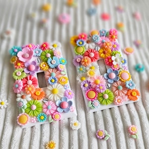May include: Two white light switch covers adorned with colorful, dimensional flowers in shades of pink, yellow, orange, blue, and green. The floral embellishments create a vibrant, textured surface. Scattered flowers are also visible.