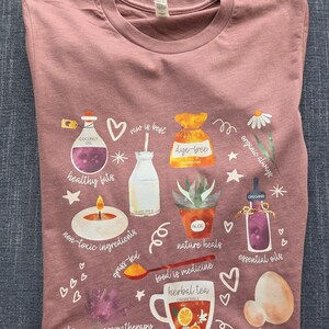 May include: A pink t-shirt with a watercolor design featuring various healthy lifestyle elements, including coconut oil, dye-free food, organic ingredients, essential oils, herbal tea, and pasture-raised products. The text on the shirt reads "raw is best", "healthy fats", "non-toxic ingredients", "grass-fed", "organic always", "dye-free", "nature heals", "food is medicine", "essential oils", "aromatherapy", and "pasture-raised".