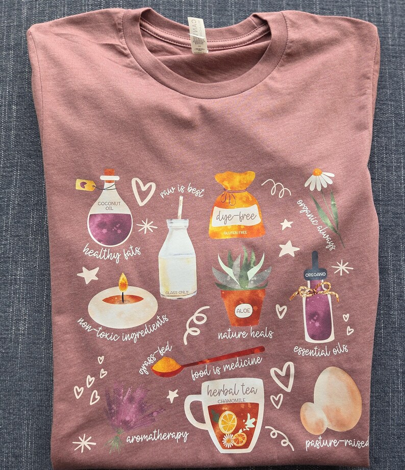 May include: A pink t-shirt with a watercolor design featuring various healthy lifestyle elements, including coconut oil, dye-free food, organic ingredients, essential oils, herbal tea, and pasture-raised products. The text on the shirt reads "raw is best", "healthy fats", "non-toxic ingredients", "grass-fed", "organic always", "dye-free", "nature heals", "food is medicine", "essential oils", "aromatherapy", and "pasture-raised".
