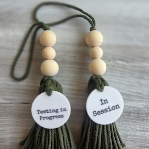 Puede incluir: Dos adornos colgantes decorativos con borlas de algod&oacute;n verde oliva y tres cuentas de madera clara natural cada uno. A cada borla se adjunta un disco circular blanco con texto negro. Un disco dice Testing in Progress y el otro dice In Session.