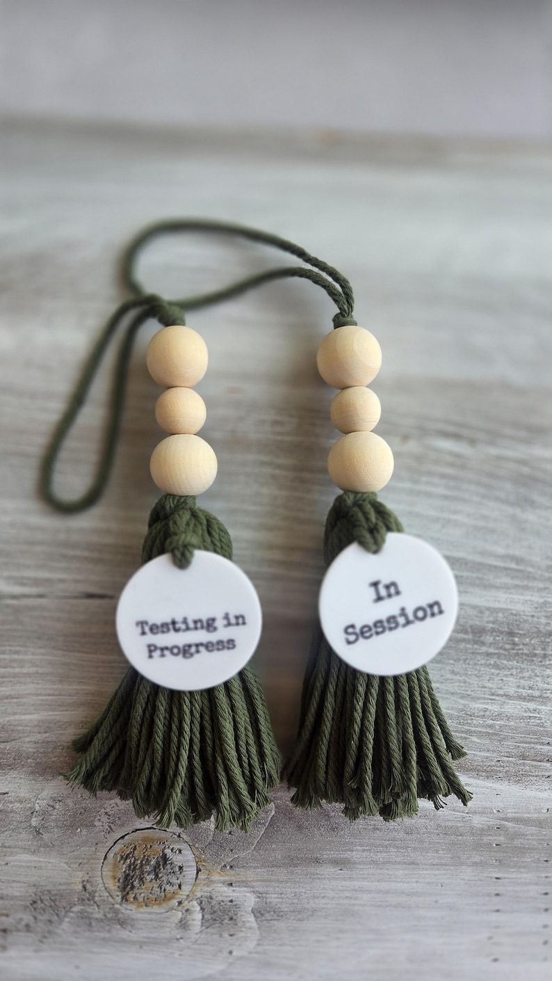 Puede incluir: Dos adornos colgantes decorativos con borlas de algod&oacute;n verde oliva y tres cuentas de madera clara natural cada uno. A cada borla se adjunta un disco circular blanco con texto negro. Un disco dice Testing in Progress y el otro dice In Session.