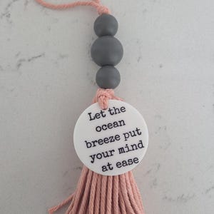 Puede incluir: Un adorno colgante decorativo que presenta un lazo de cuerda color melocot&oacute;n, tres cuentas esf&eacute;ricas de color gris mate y un disco circular blanco impreso con el texto negro Let the ocean breeze put your mind at ease, acabado con una borla.