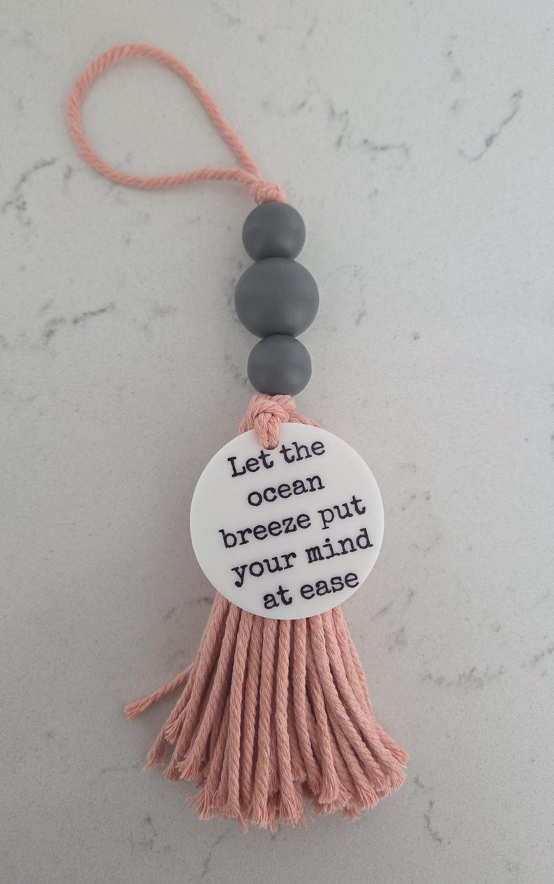 Puede incluir: Un adorno colgante decorativo que presenta un lazo de cuerda color melocot&oacute;n, tres cuentas esf&eacute;ricas de color gris mate y un disco circular blanco impreso con el texto negro Let the ocean breeze put your mind at ease, acabado con una borla.