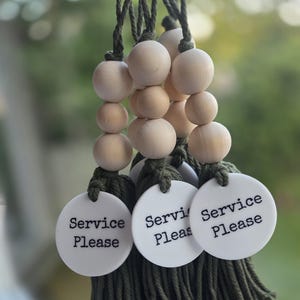 Puede incluir: Tres adornos colgantes decorativos con borlas de cord&oacute;n verde oliva y cuerdas de cuentas de madera natural de color claro. Cada adorno incluye un disco redondo de acr&iacute;lico blanco impreso con el texto negro Service Please.