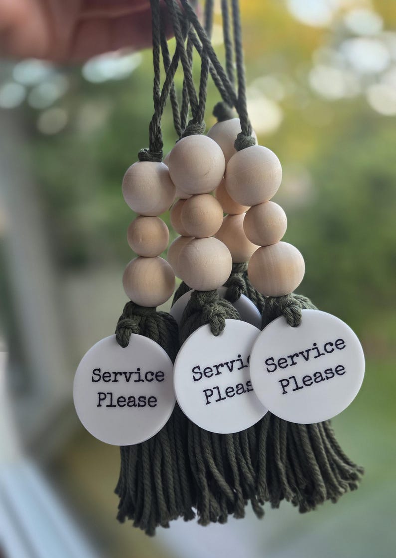 Puede incluir: Tres adornos colgantes decorativos con borlas de cord&oacute;n verde oliva y cuerdas de cuentas de madera natural de color claro. Cada adorno incluye un disco redondo de acr&iacute;lico blanco impreso con el texto negro Service Please.