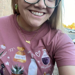 May include: A pink t-shirt with a graphic print of various items, including a bag labeled "sage-free", a bottle of milk, a bottle of purple liquid labeled "tumojoj oil", a candle, and a potted succulent. The text "self-love" is printed on the bag. The text "good!" is printed above the heart. The text "little rituals" is printed below the bottle of purple liquid. The text "self-love" is printed below the potted succulent. The text "lail" is printed below the candle. The text "and it" is printed below the bottle of milk. The text "os-gub" is printed above the bag. The text "30 JA" is printed on the pot. The text "tan" is printed below the pot. The text "wat li" is printed below the candle. The text "Crowon" is printed above the bag. The text "ne" is printed below the bottle of purple liquid. The text "огрел сига" is printed above the bag. The text "A" is printed below the bottle of milk. The text "لا" is printed below the candle.