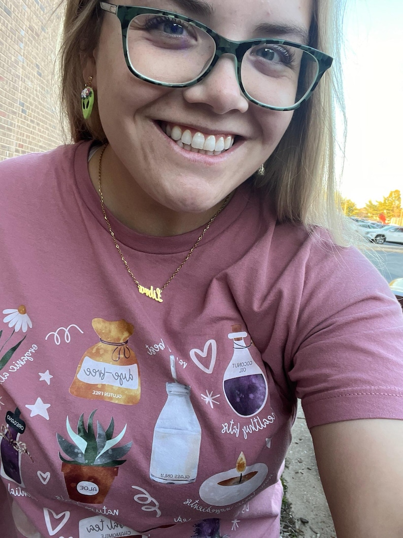 May include: A pink t-shirt with a graphic print of various items, including a bag labeled "sage-free", a bottle of milk, a bottle of purple liquid labeled "tumojoj oil", a candle, and a potted succulent. The text "self-love" is printed on the bag. The text "good!" is printed above the heart. The text "little rituals" is printed below the bottle of purple liquid. The text "self-love" is printed below the potted succulent. The text "lail" is printed below the candle. The text "and it" is printed below the bottle of milk. The text "os-gub" is printed above the bag. The text "30 JA" is printed on the pot. The text "tan" is printed below the pot. The text "wat li" is printed below the candle. The text "Crowon" is printed above the bag. The text "ne" is printed below the bottle of purple liquid. The text "огрел сига" is printed above the bag. The text "A" is printed below the bottle of milk. The text "لا" is printed below the candle.