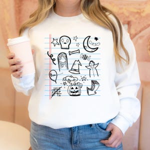 May include: White sweatshirt with a black and white graphic print of Halloween doodles on a lined notebook page. The doodles include a skull, a ghost, a witch's hat, a vampire, a pumpkin, a crescent moon, a bat, a candy cane, a bone, and the word "Boo!"