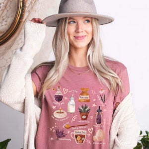 May include: A pink t-shirt with a graphic design featuring various illustrations related to natural wellness. The illustrations include a bottle of essential oil, a bag of dye-free tea, a candle, a jar of essential oil, a potted succulent, a glass of milk, a cup of herbal tea, and an egg. The text on the shirt includes "healthy fats", "non-toxic ingredients", "dye-free", "nature heals", "food is medicine", "essential oils", "aromatherapy", and "pasture raised".