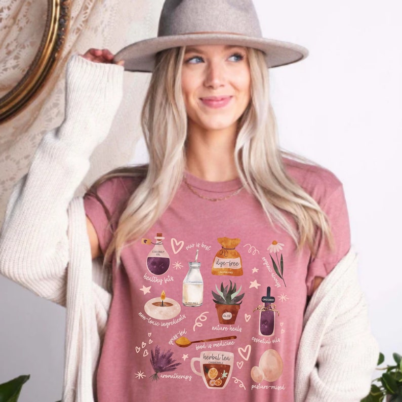 May include: A pink t-shirt with a graphic design featuring various illustrations related to natural wellness. The illustrations include a bottle of essential oil, a bag of dye-free tea, a candle, a jar of essential oil, a potted succulent, a glass of milk, a cup of herbal tea, and an egg. The text on the shirt includes "healthy fats", "non-toxic ingredients", "dye-free", "nature heals", "food is medicine", "essential oils", "aromatherapy", and "pasture raised".
