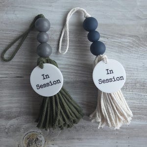 Puede incluir: Dos letreros colgantes para puerta con un disco circular blanco y el texto negro In Session. Uno tiene tres cuentas de madera gris y una borla de algod&oacute;n verde oliva con un lazo a juego. El otro tiene tres cuentas azul marino y una borla crema.