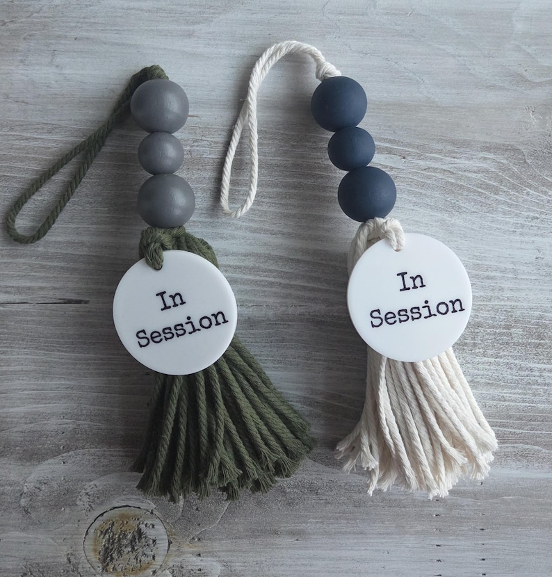Puede incluir: Dos letreros colgantes para puerta con un disco circular blanco y el texto negro In Session. Uno tiene tres cuentas de madera gris y una borla de algod&oacute;n verde oliva con un lazo a juego. El otro tiene tres cuentas azul marino y una borla crema.