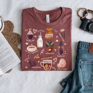May include: A maroon t-shirt with a white graphic design featuring various illustrations related to a healthy lifestyle. The illustrations include a bottle of coconut oil, a glass of milk, a potted aloe vera plant, a cup of herbal tea, and a jar of essential oils. The text on the shirt includes "raw is best", "dye-free", "organic always", "healthy fats", "non-toxic ingredients", "nature heals", "food is medicine", "essential oils", "aromatherapy", and "pasture-raised".