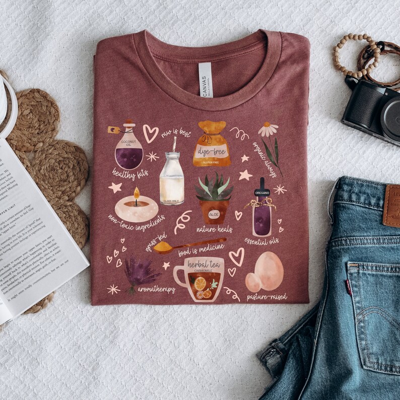 May include: A maroon t-shirt with a white graphic design featuring various illustrations related to a healthy lifestyle. The illustrations include a bottle of coconut oil, a glass of milk, a potted aloe vera plant, a cup of herbal tea, and a jar of essential oils. The text on the shirt includes "raw is best", "dye-free", "organic always", "healthy fats", "non-toxic ingredients", "nature heals", "food is medicine", "essential oils", "aromatherapy", and "pasture-raised".