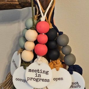 Puede incluir: Una colecci&oacute;n de letreros colgantes para puertas con cuentas de madera redondas en varios colores, incluyendo rojo, negro, gris y crema. Cada letrero tiene un disco circular blanco con texto negro que dice meeting in progress, open, occupied, privacy o not disturb.