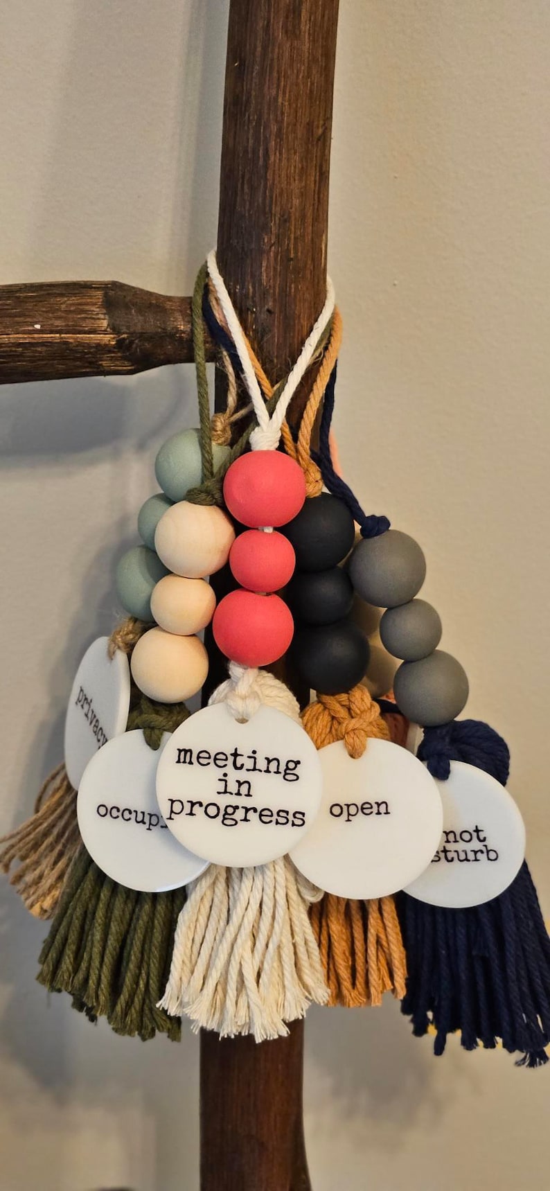 Puede incluir: Una colecci&oacute;n de letreros colgantes para puertas con cuentas de madera redondas en varios colores, incluyendo rojo, negro, gris y crema. Cada letrero tiene un disco circular blanco con texto negro que dice meeting in progress, open, occupied, privacy o not disturb.