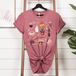 May include: A pink t-shirt with a graphic design featuring various illustrations related to natural wellness. The design includes a bottle of healthy fats, a jar of lye-free soap, a candle, a cup of herbal tea, a jar of essential oils, and a bag of nature-raised eggs. The text on the shirt reads "* is best", "lye-free", "healthy fats", "aromatic ingredients", "nature heals", "food is medicine", "herbal tea", "aromatherapy", and "pasture-raised".