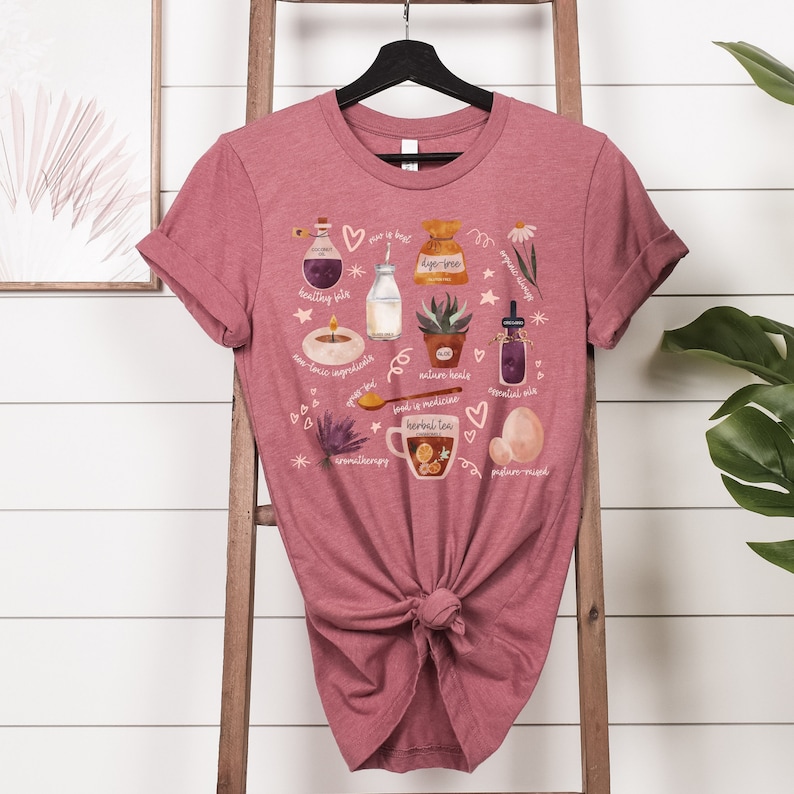 May include: A pink t-shirt with a graphic design featuring various illustrations related to natural wellness. The design includes a bottle of healthy fats, a jar of lye-free soap, a candle, a cup of herbal tea, a jar of essential oils, and a bag of nature-raised eggs. The text on the shirt reads "* is best", "lye-free", "healthy fats", "aromatic ingredients", "nature heals", "food is medicine", "herbal tea", "aromatherapy", and "pasture-raised".