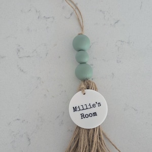 Puede incluir: Un adorno colgante decorativo que presenta tres cuentas de color verde salvia, un disco circular de acr&iacute;lico blanco con el texto impreso en negro Millie's Room y una borla de hilo de yute natural, suspendido de un lazo de yute a juego.