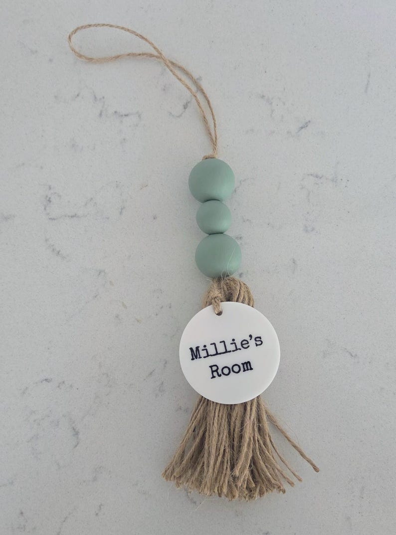 Puede incluir: Un adorno colgante decorativo que presenta tres cuentas de color verde salvia, un disco circular de acr&iacute;lico blanco con el texto impreso en negro Millie's Room y una borla de hilo de yute natural, suspendido de un lazo de yute a juego.