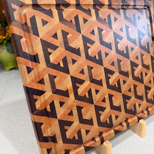May include: A rectangular wooden cutting board featuring an intricate geometric pattern composed of light, medium, and dark brown wood segments. The surface displays a repeating three-dimensional cube design with a carved juice groove along the edge.