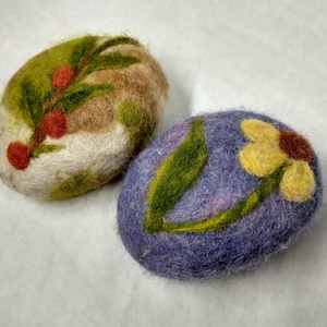 May include: Two oval-shaped felted soap bars. One is a mix of green, brown, and white with a red berry branch design. The other is purple with a yellow flower and green leaf design. Handmade soap.