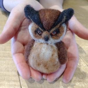 May include: A small, brown and black felt owl with large, orange eyes. The owl is sitting in the palm of a person's hand.