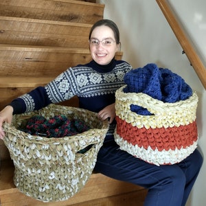 May include: Two crocheted baskets made from recycled fabric. The larger basket is beige and white with a green, red, and black patterned blanket inside. The smaller basket is white, yellow, red, and blue. A person is sitting on stairs with the baskets in front of them.