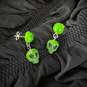Op de afbeelding: Een paar groene alien schedel oorhangers met een groene bol aan de bovenkant van elke oorhanger.